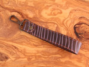 Schlüsselanhänger mit schwarzen Karabiner, bemalt mit Streifen in lila, umnäht in den Garnfarben orange und flieder