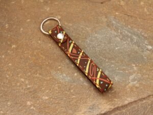 Kleiner Schlüsselanhänger aus Leder in mittelbraun mit Muster und Streifen in gelb und orange. Die Garnfarbe ist rot und gelb.